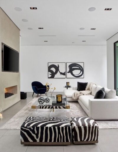 Modern living room with minimalist decor, featuring neutral colors, a zebra-patterned ottoman, and large windows.