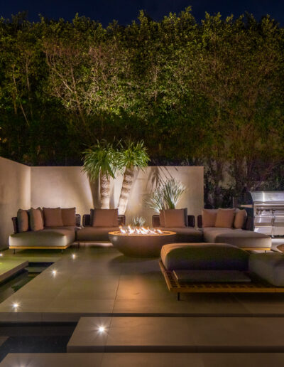 An outdoor seating area at night featuring modern furniture, a fire pit, ambient lighting, and lush greenery.