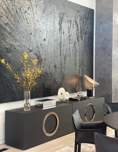 A modern room with minimalist furniture featuring a large abstract black painting, a console table with decorative objects, and a round table with black chairs.